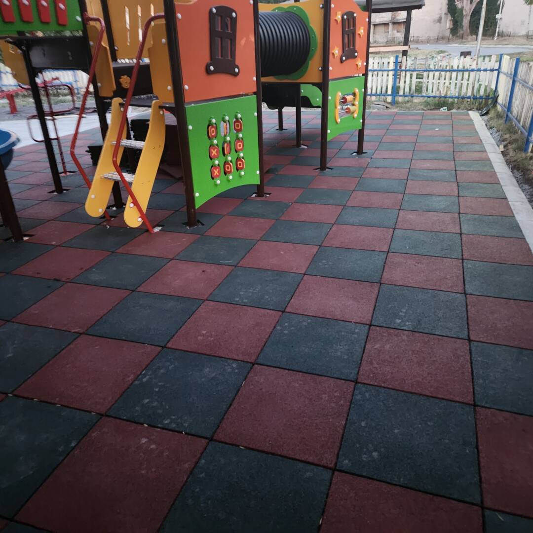 montaj-kauchukova-nastilka-detska-ploshtadka-tereno.jpg Installed "Tereno Eco" shock-absorbing rubber flooring in a chessboard pattern (red and black) under playground equipment.